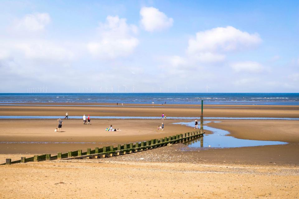 Rhyl beach on a sunny morning with people and wind turbines on the horizon.