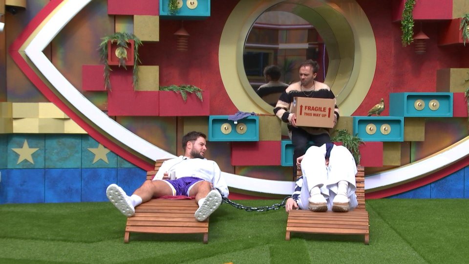 Three men on a reality TV show; two are chained together on lounge chairs, while the third holds a box.