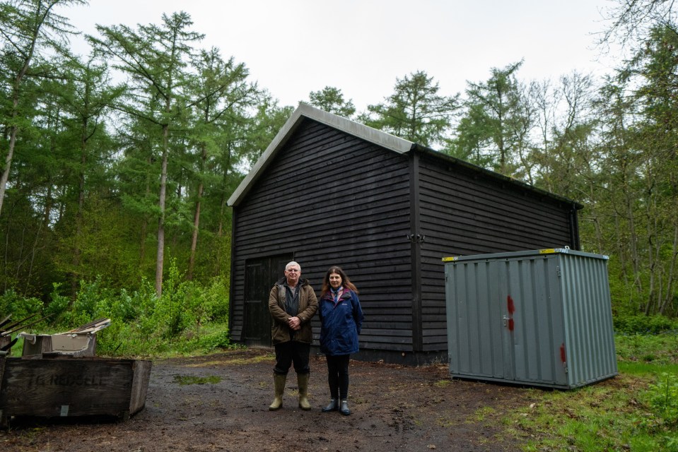 A man and woman stand in front of a dark wooden building in a wooded area.