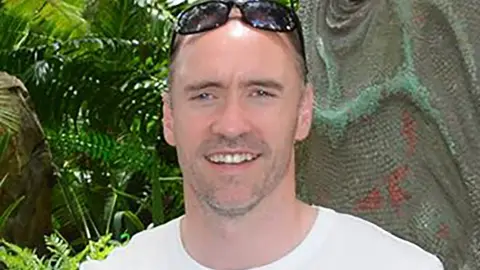 BBC Paul Doyle has short dark greying hair with sunglasses on his head and is wearing a white T-shirt.