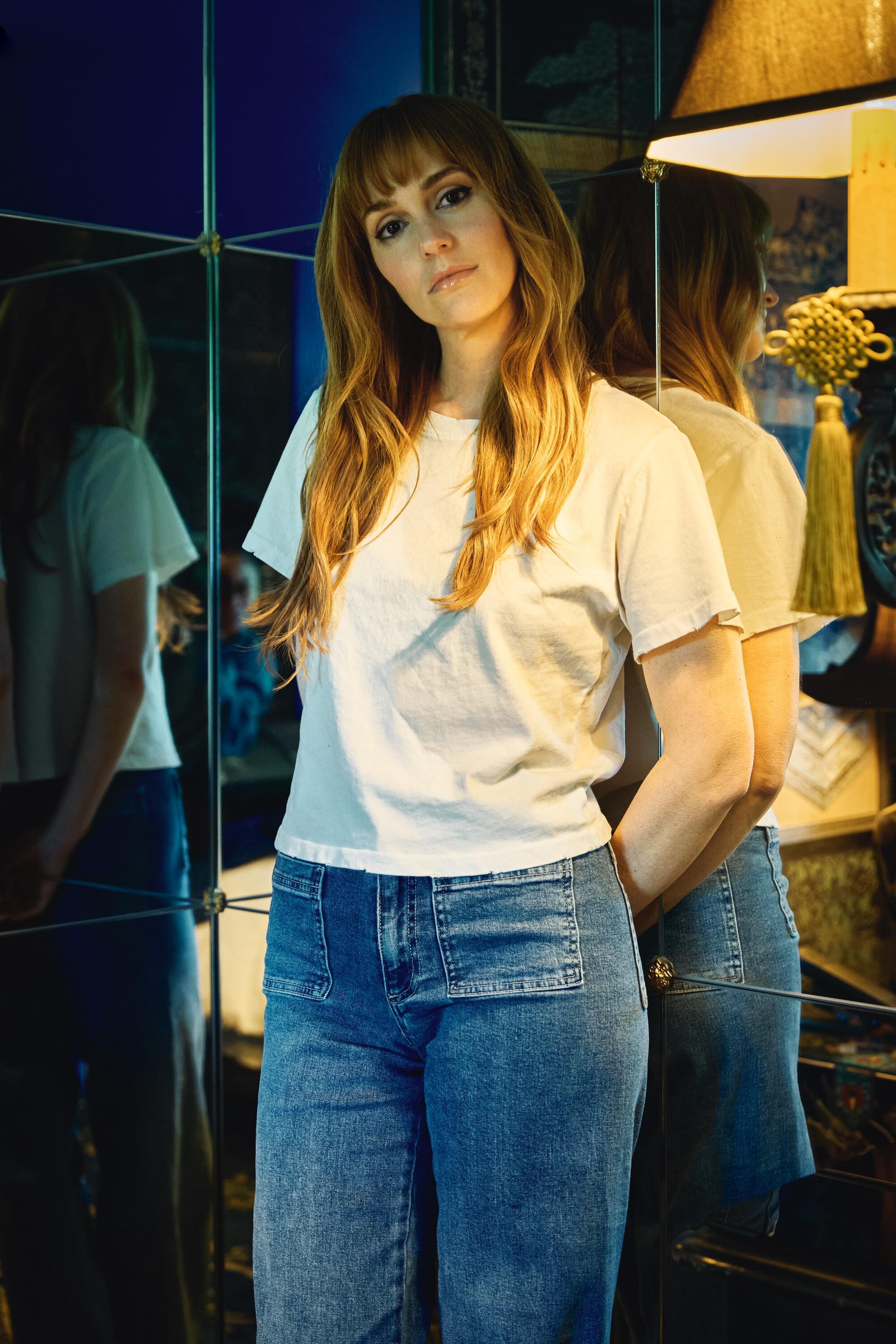 A woman in white T-shirt and blue jeans stands against a mirror, hands behind her, head tilted