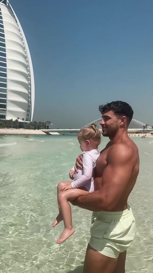 Man holding a toddler in shallow water with a tall building in the background.