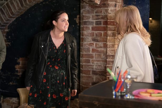Jacqueline Jossa in a black dress and jacket, stood beside Michelle Collins in a white jacket, in a still from EastEnders.