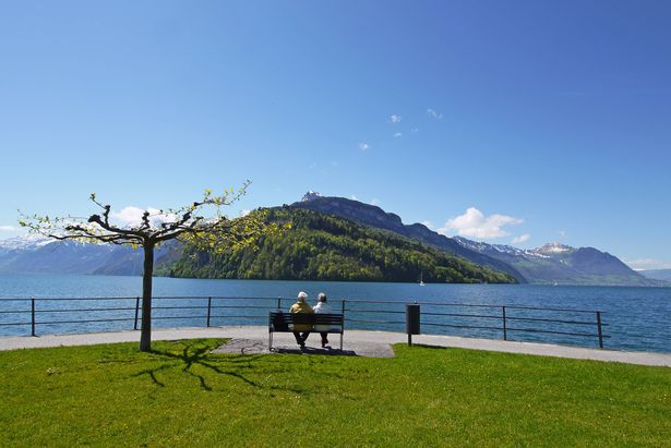 Swiss couple in their seventies sitting on bench admiring Lake Lucerne relaxing. Great mountain scenery in springtime. Swiss Alps, blue sky with clouds, sunshine. Lake promenade at Brunnen, Canton Schwyz, Switzerland.