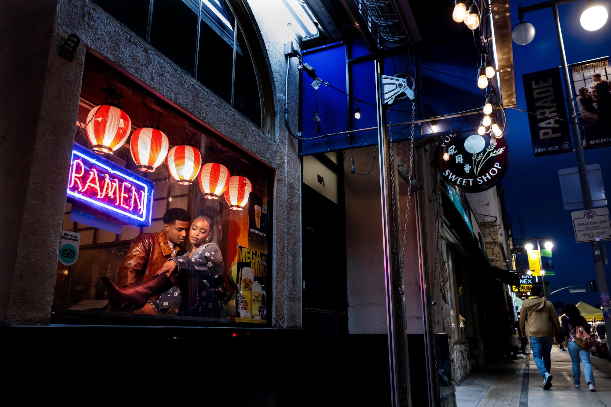 A man and a woman lean against the window of a restaurant with a glowing neon light that reads "ramen."