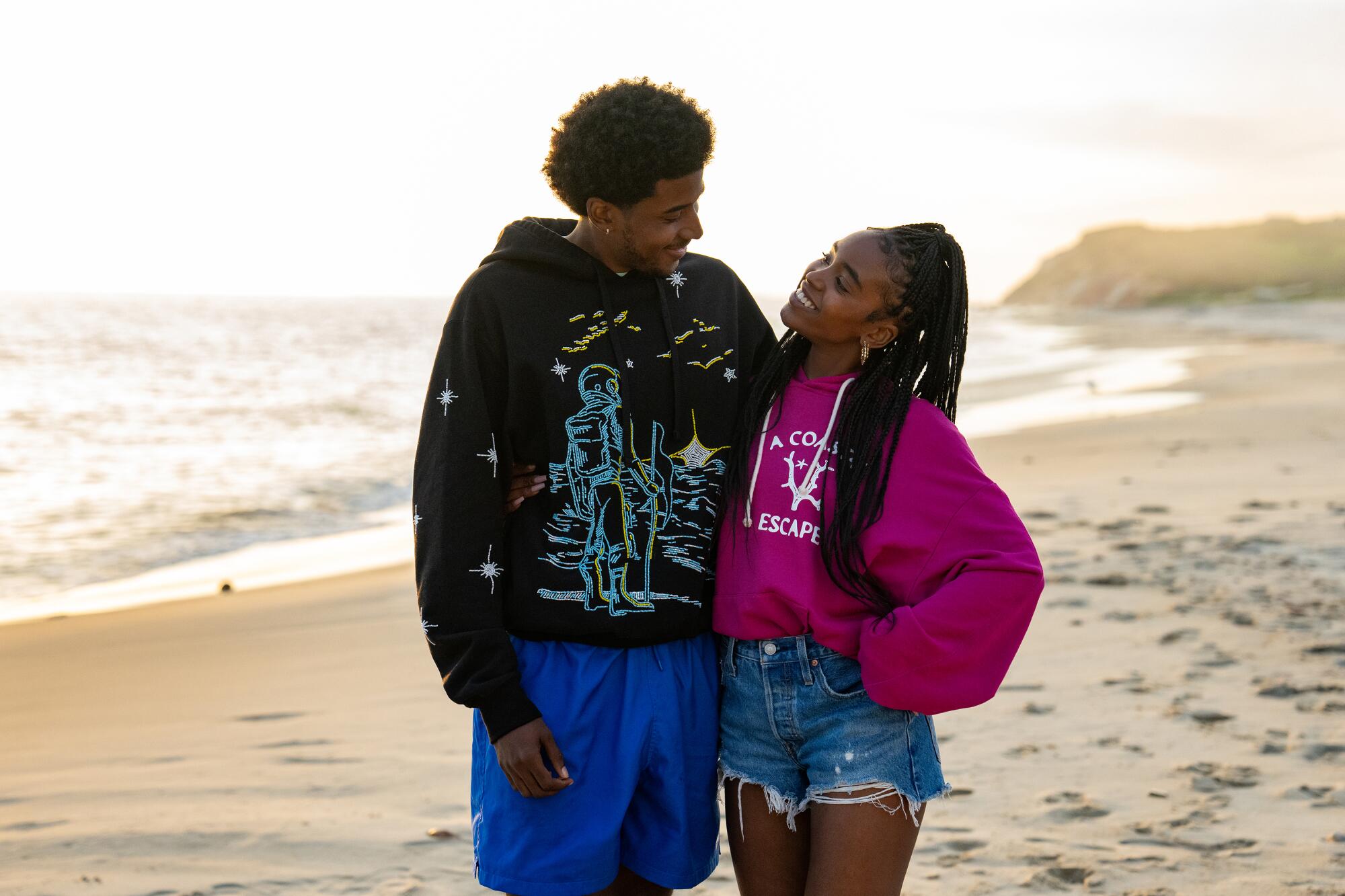 A guy in shorts and a hoodie stands besides a girl in shorts and a hoodie on a beach