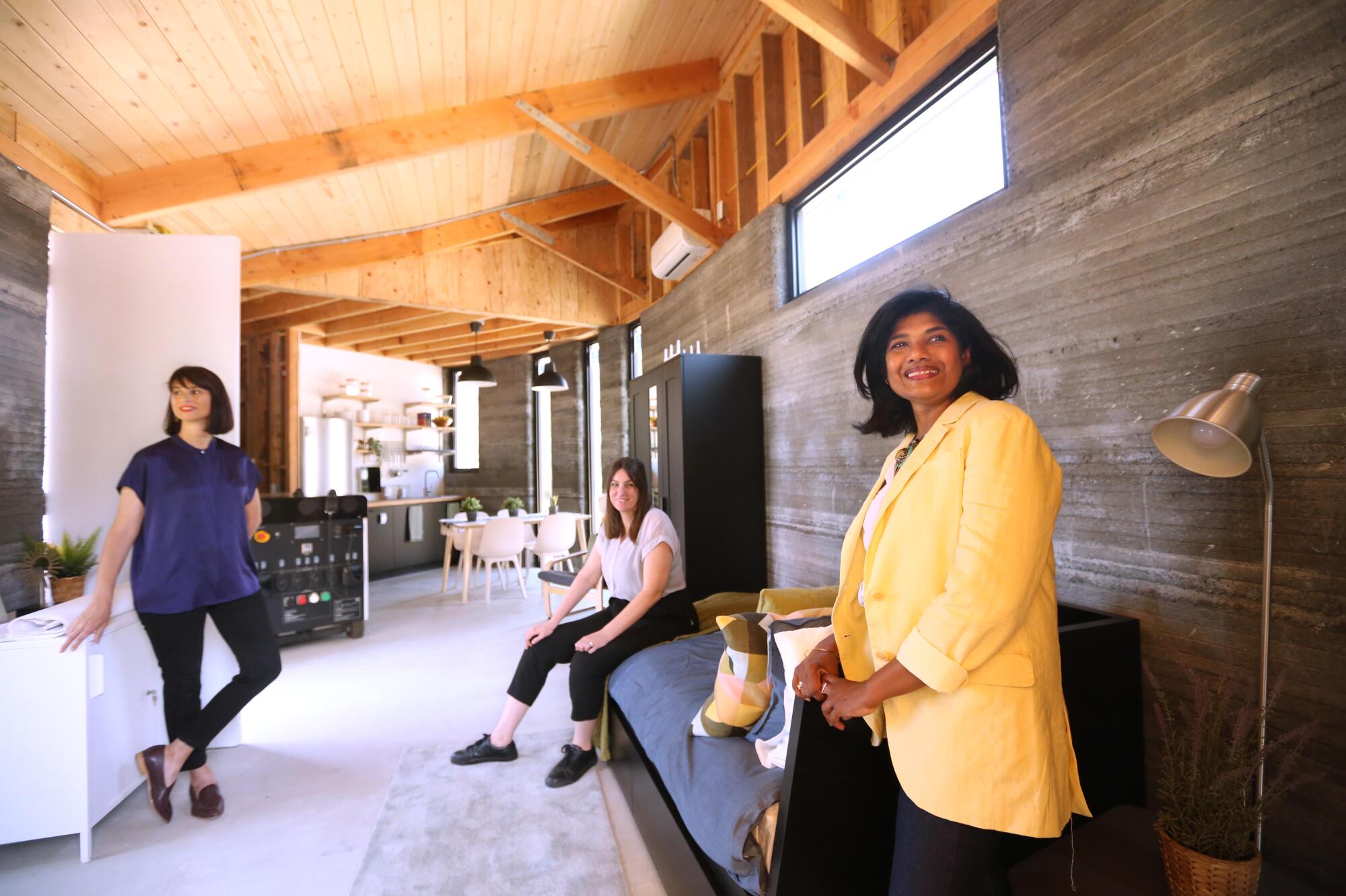 Three people sit inside a long studio with striated, 3D-printed concrete walls topped by a vaulted wood ceiling.