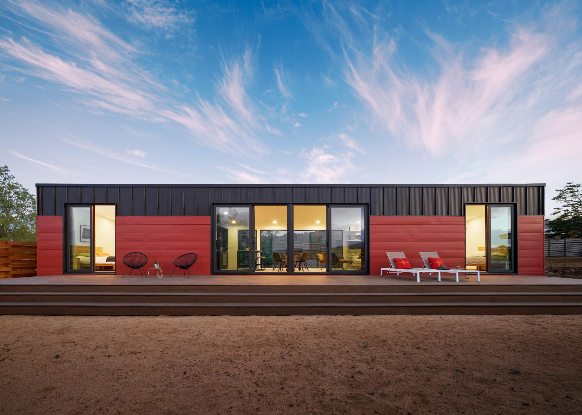 A long modular home clad in red panels sits on a desert landscape against a blue sky streaked with clouds.