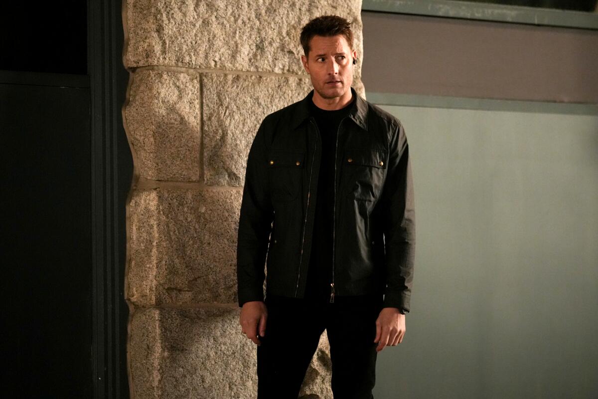 A man in a matching black ensemble stands in front of a wall