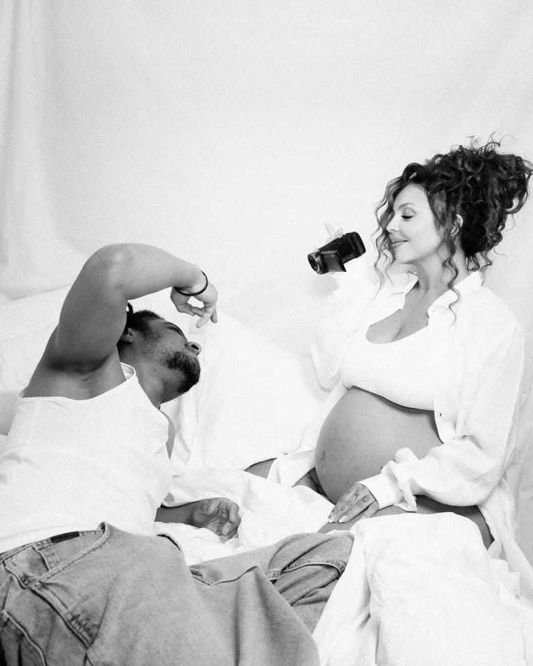 Black and white photo of a pregnant woman and a man lying in bed.  The woman holds a camcorder.