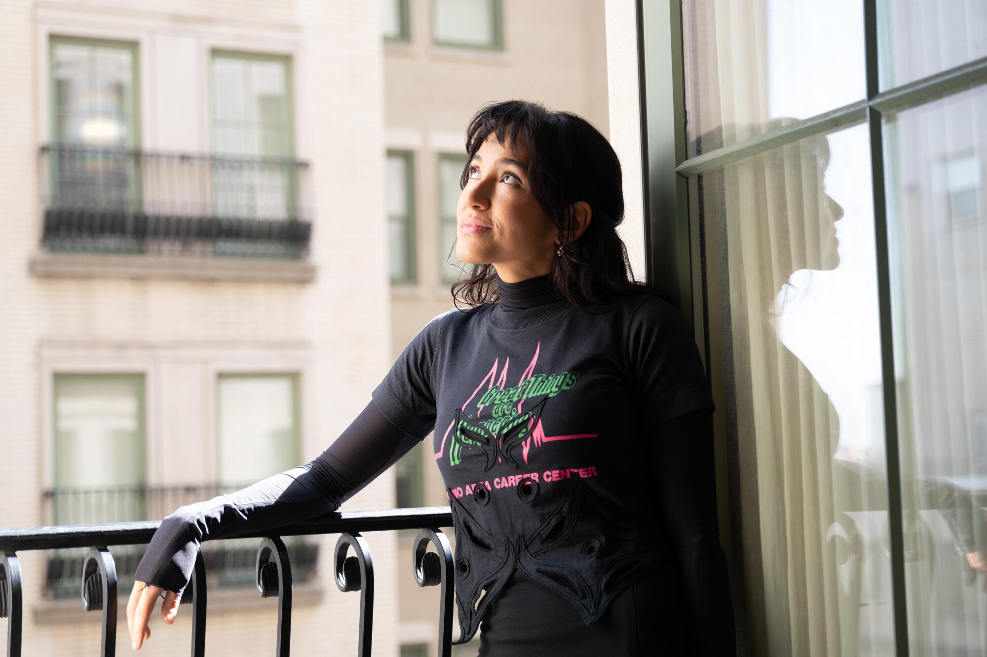 A woman with dark hair in a black long-sleeve shirt leans against the railing of an open window.