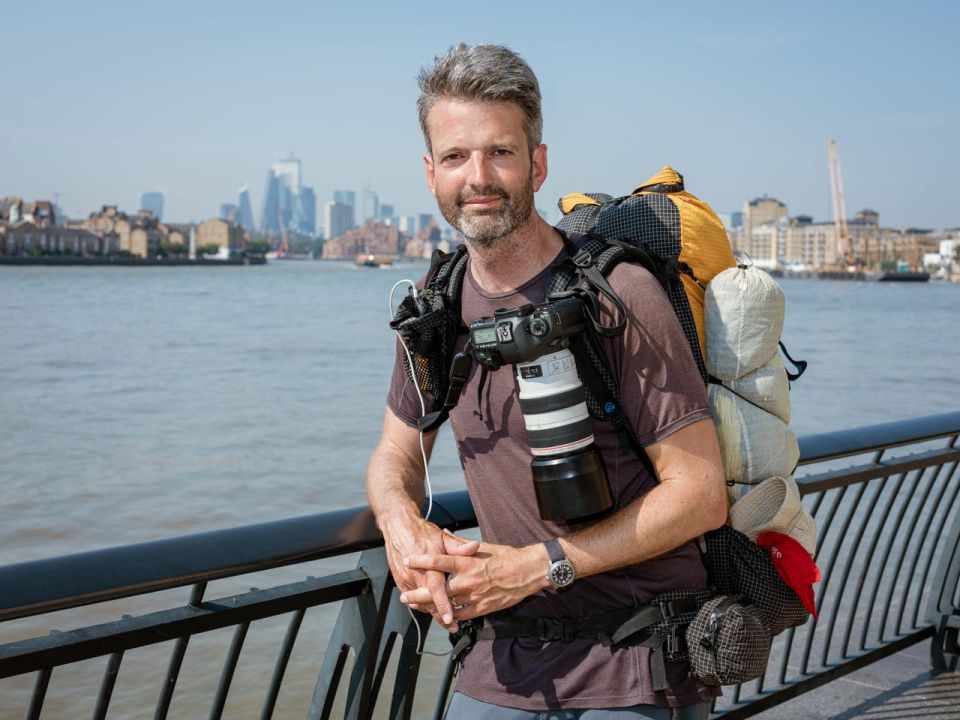 Photographer with a large backpack and camera, standing by a river with a city skyline in the background.