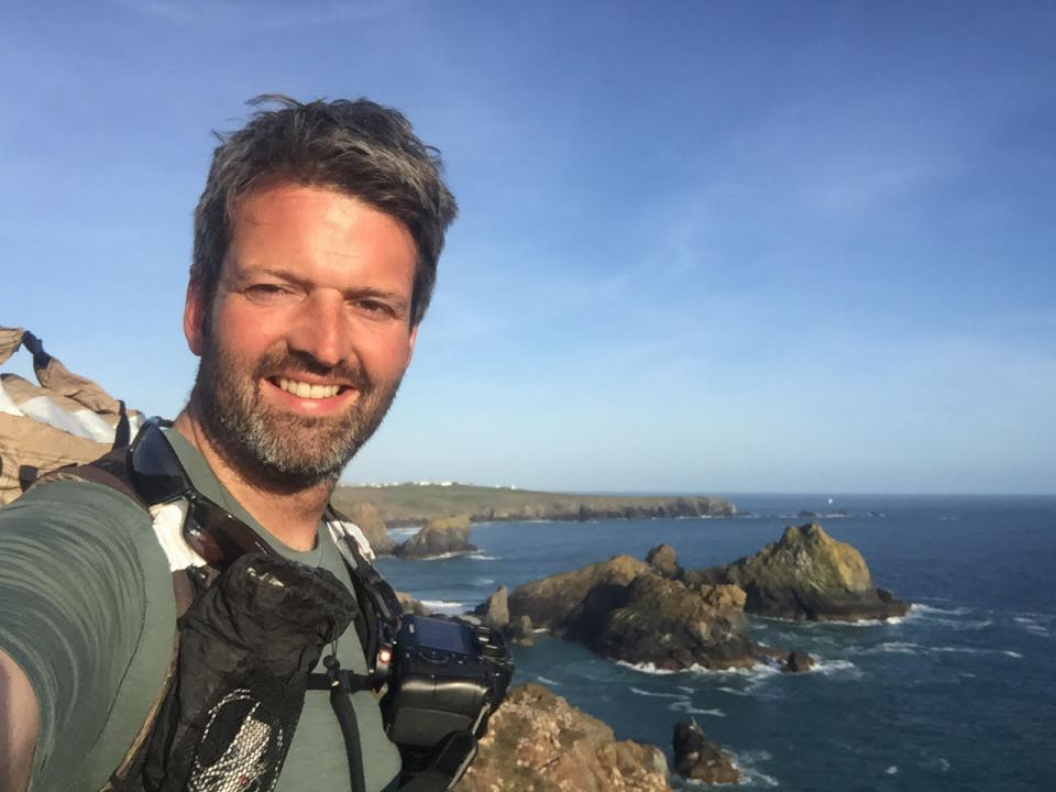 Man smiling for a selfie, wearing a backpack and camera, overlooking the ocean.