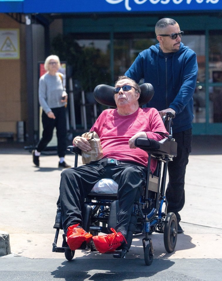 Tim Curry in a wheelchair, being pushed by another person.