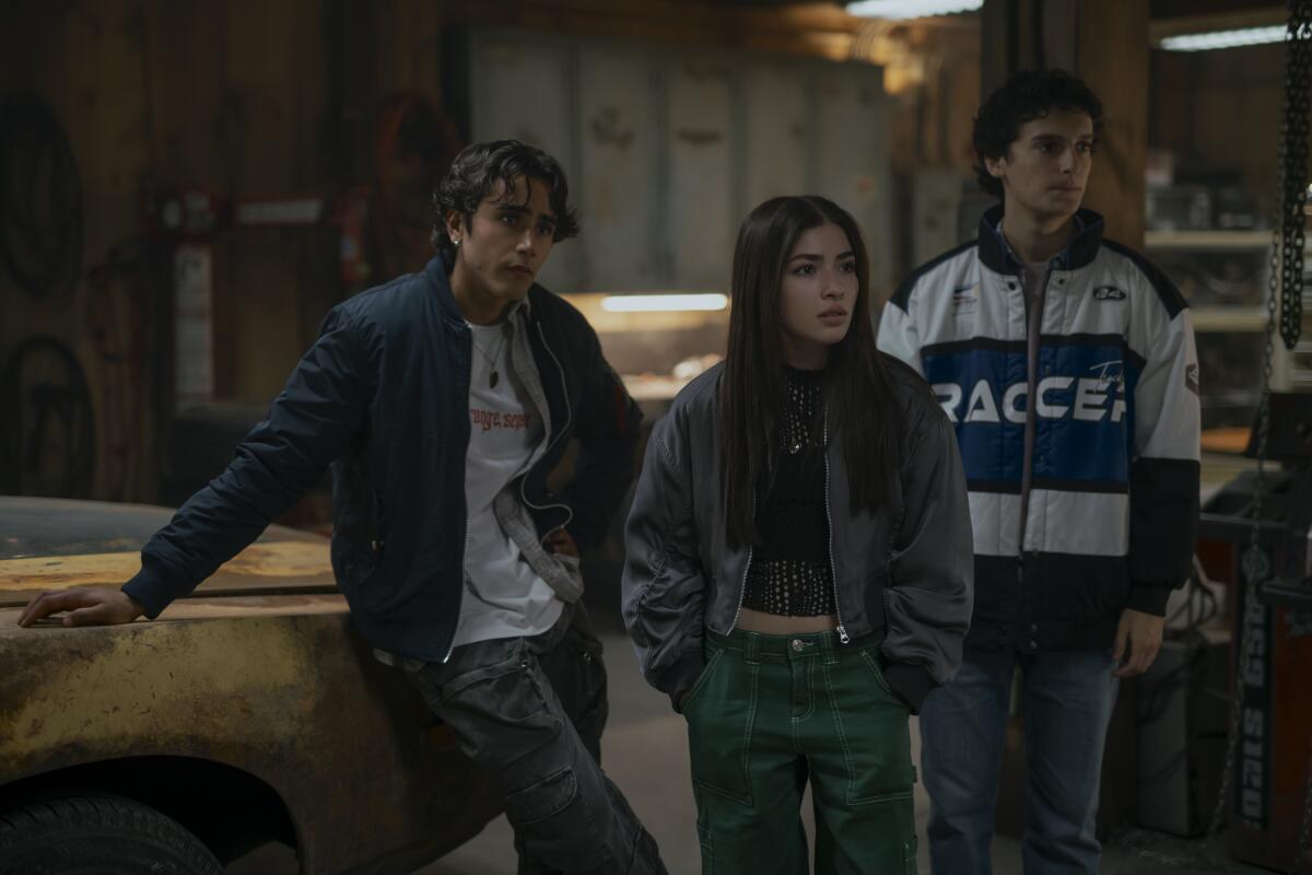 Three teenagers stand near a rusty car in a garage.