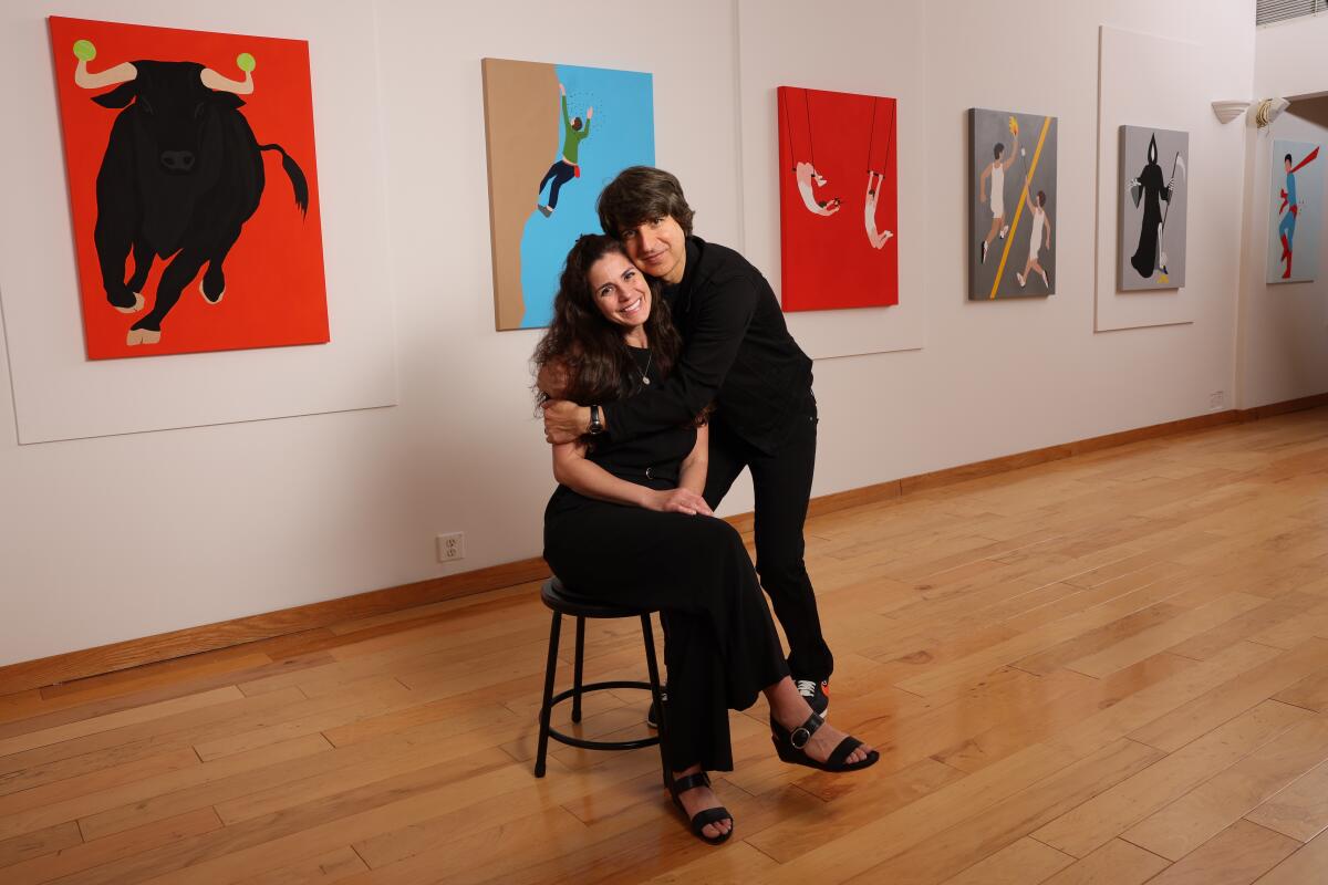 Demetri Martin hugs his wife, Rachael Beame Martin, seated on a stool in front of colorful paintings.