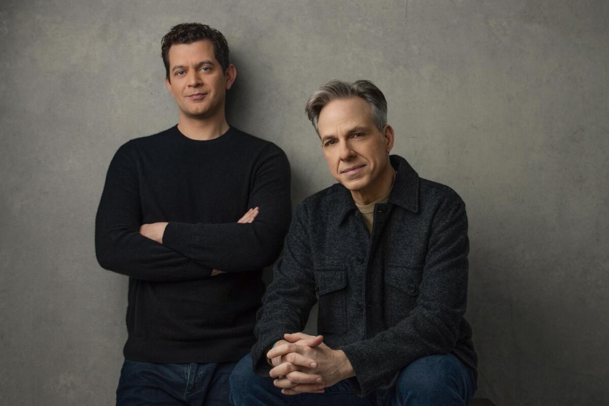 Alex Thompson stands against a wall with arms crossed while Jake Tapper sits with hands folded.