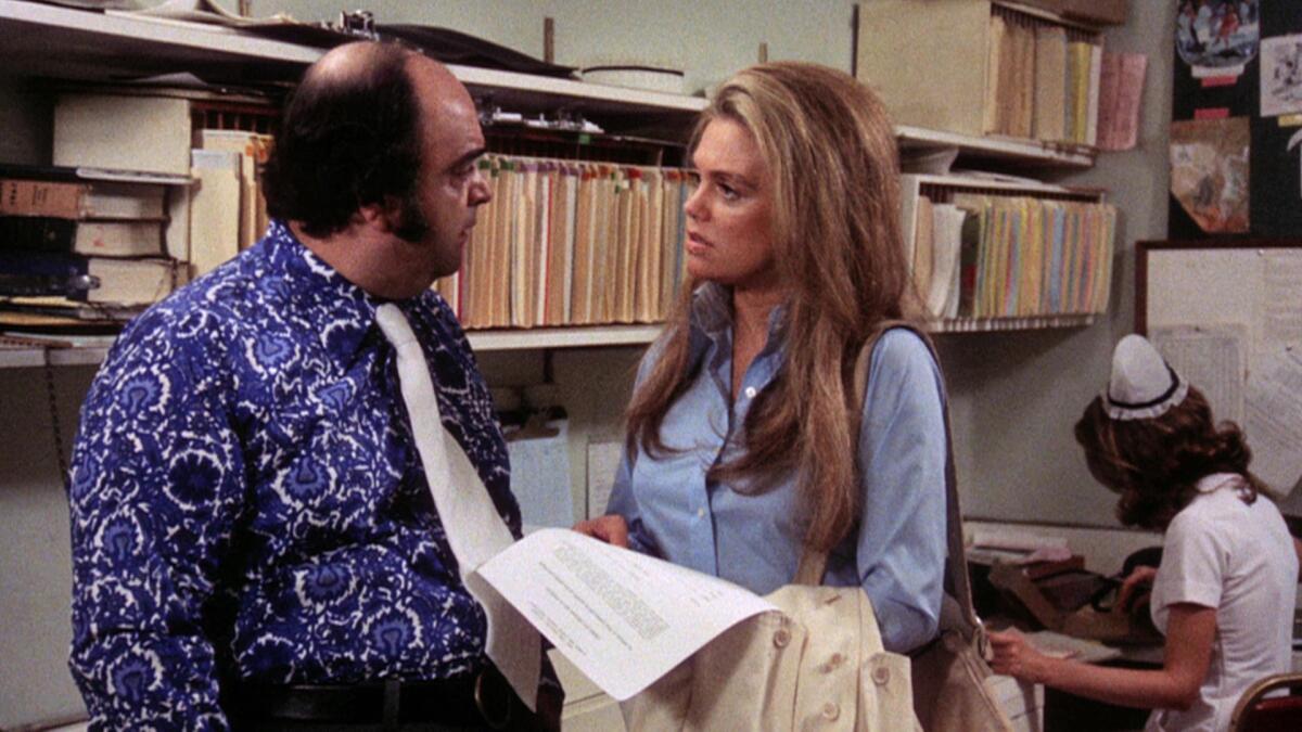 A man in a loud purple shirt and a woman have a discussion in an office.