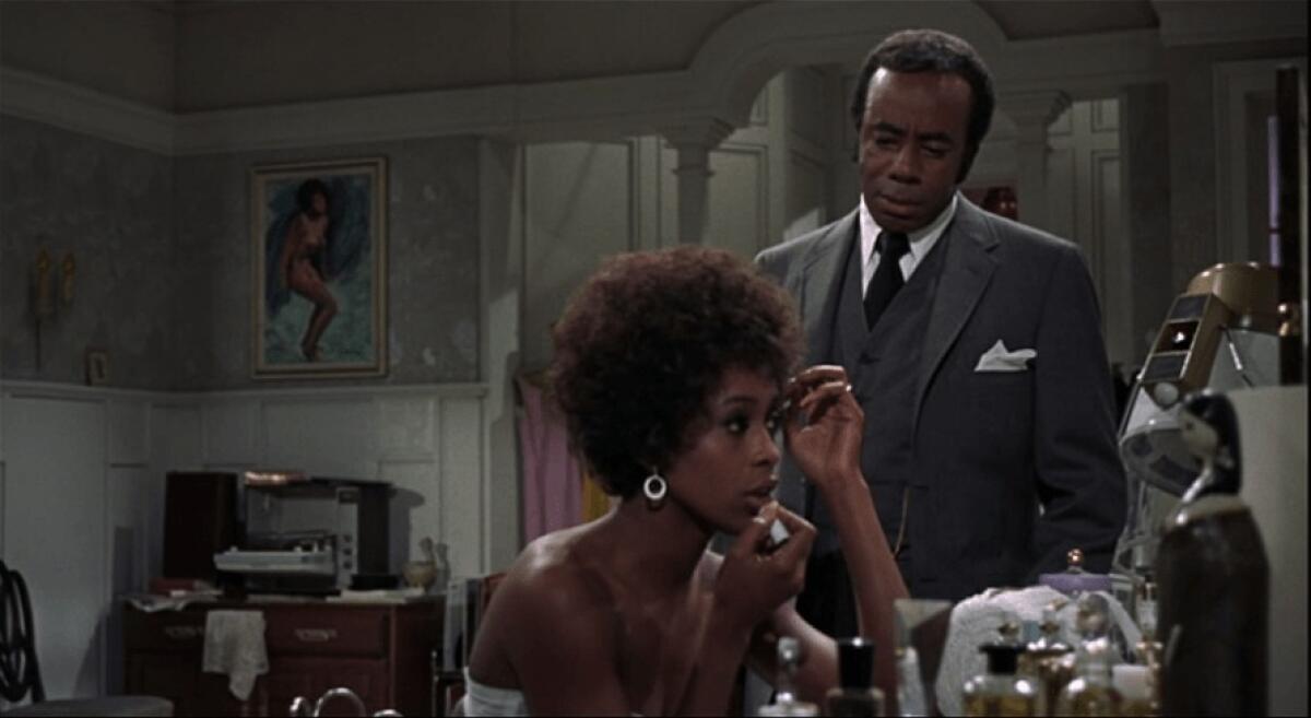 A woman puts on makeup while a man stands over her.