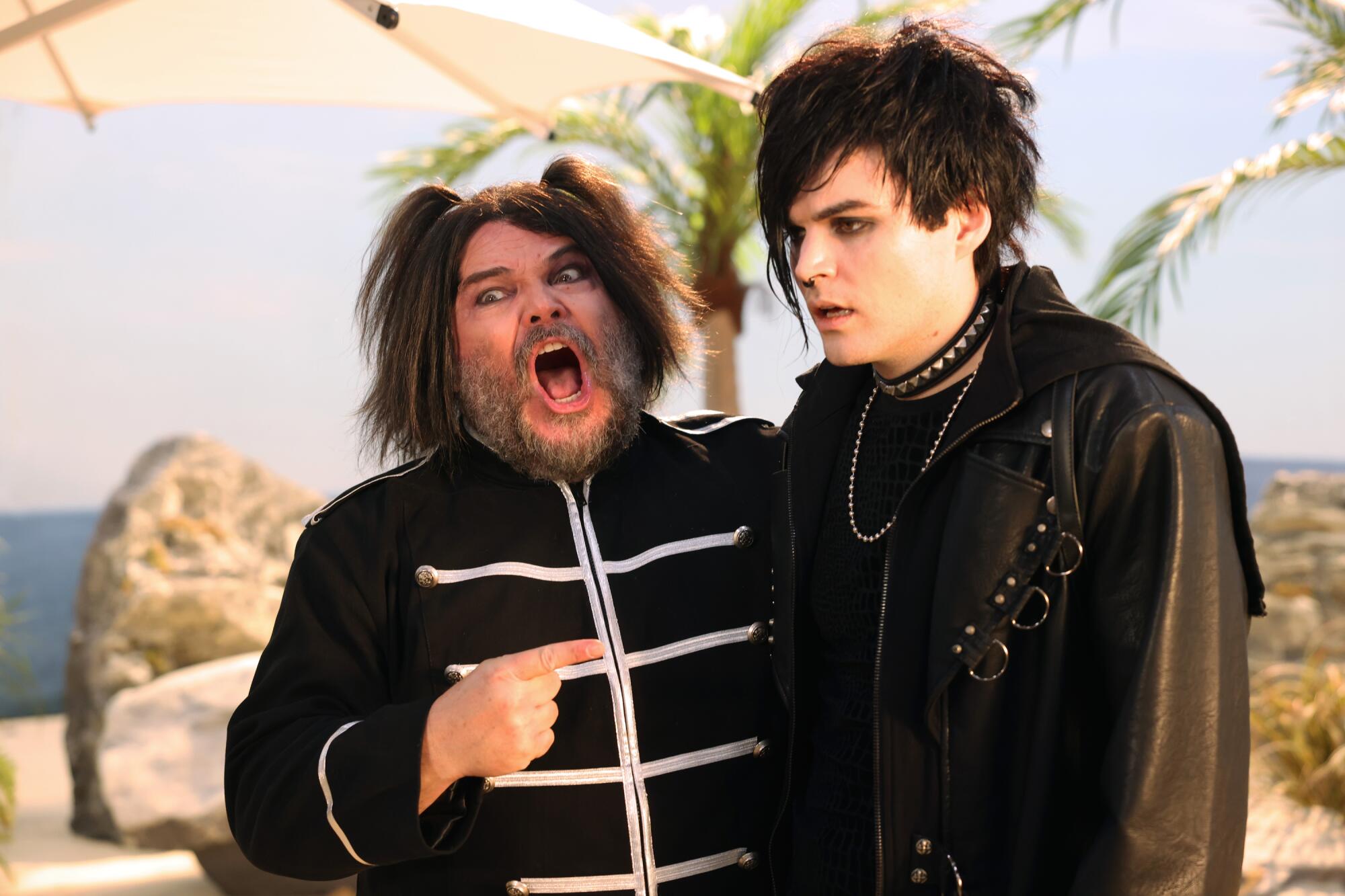 A man in a black uniform points at a teenage boy in leather jacket and black T-shirt on a beach.
