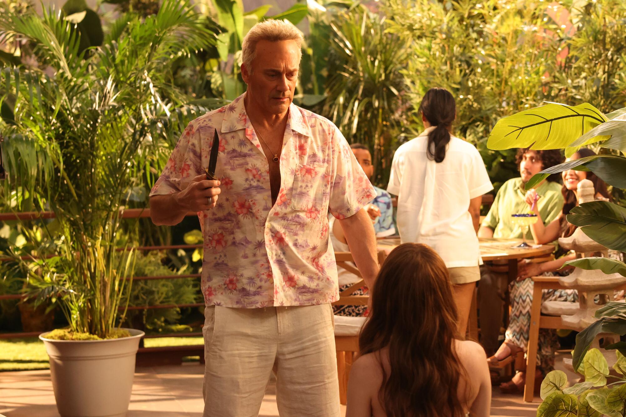 A man with white hair in a floral shirt and linen pants holds a knife as he looks at a sitting woman seen behind.