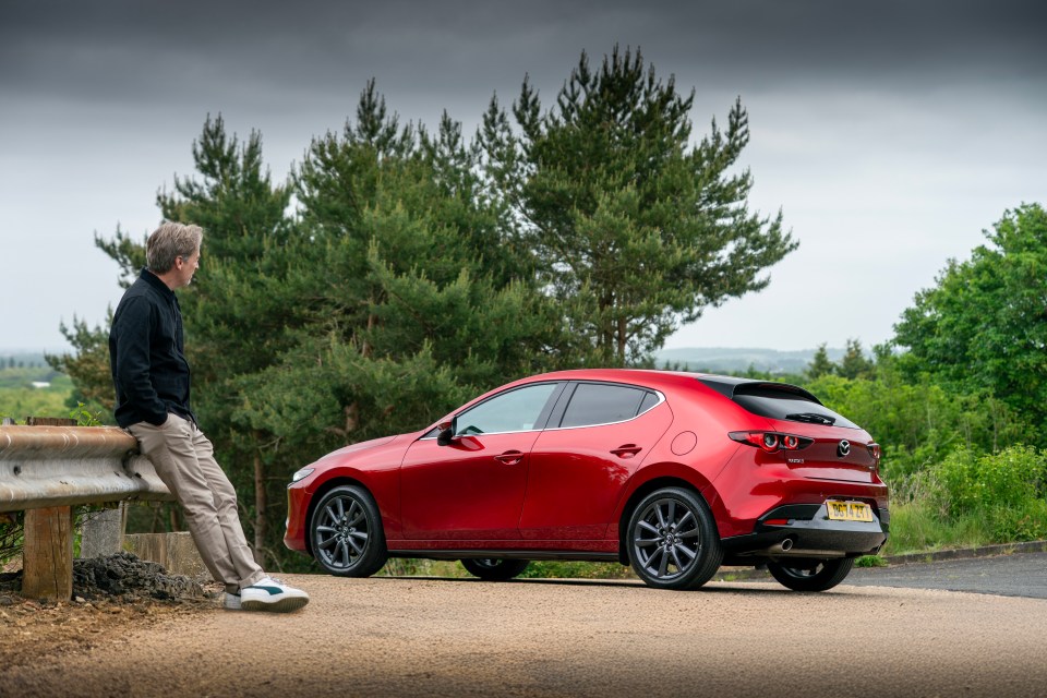 Man leaning on a barrier looking at a red Mazda 3.