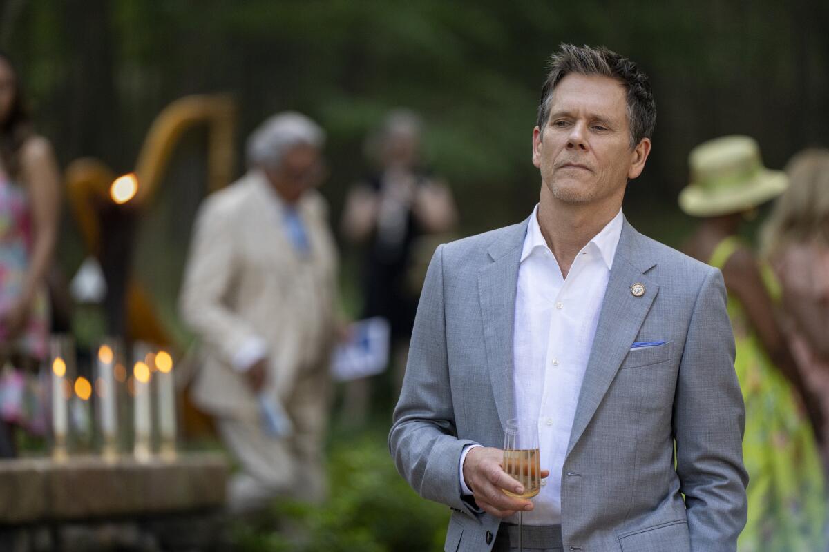 Kevin Bacon in a gray suit and white shirt holds a champagne flute in one hand, his eyes cast to the side.