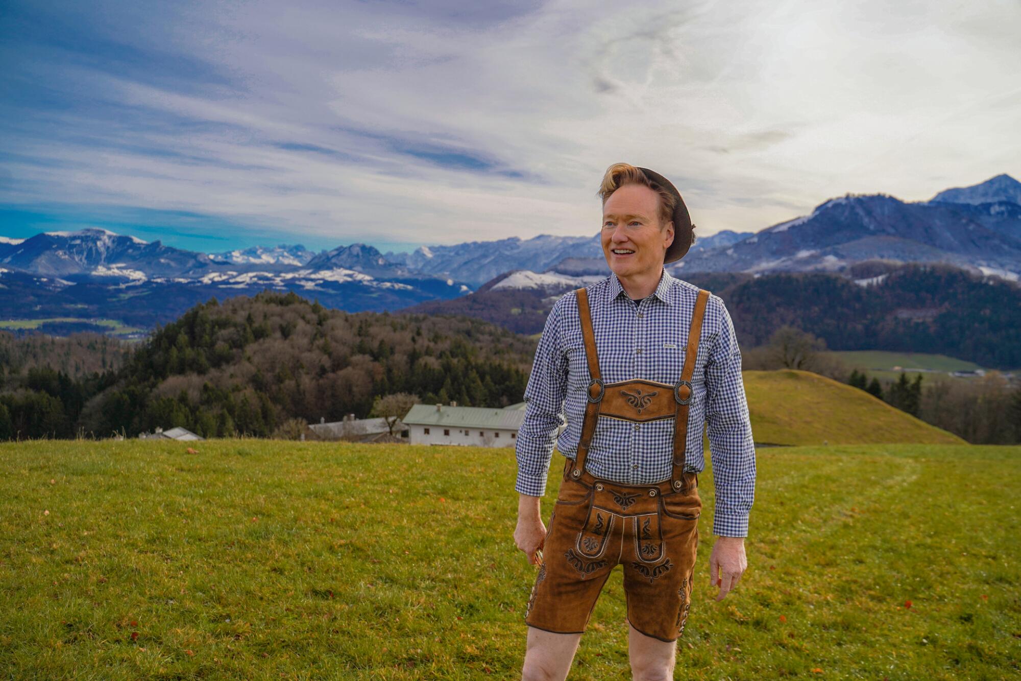 A man in lederhosen, traditional German clothing, stands on a hilltop.