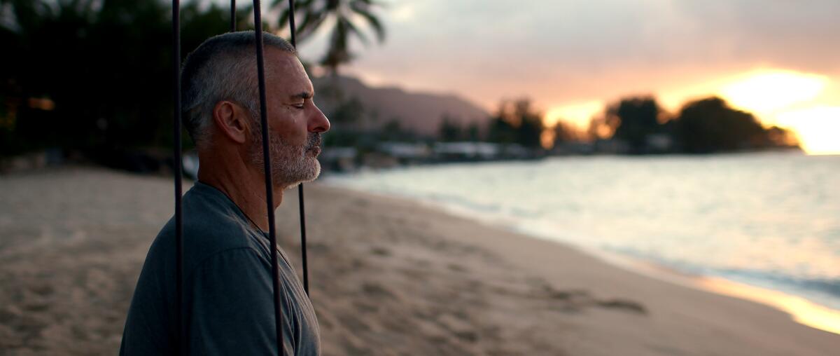 A man sits on a beach, facing the water, with his eyes closes