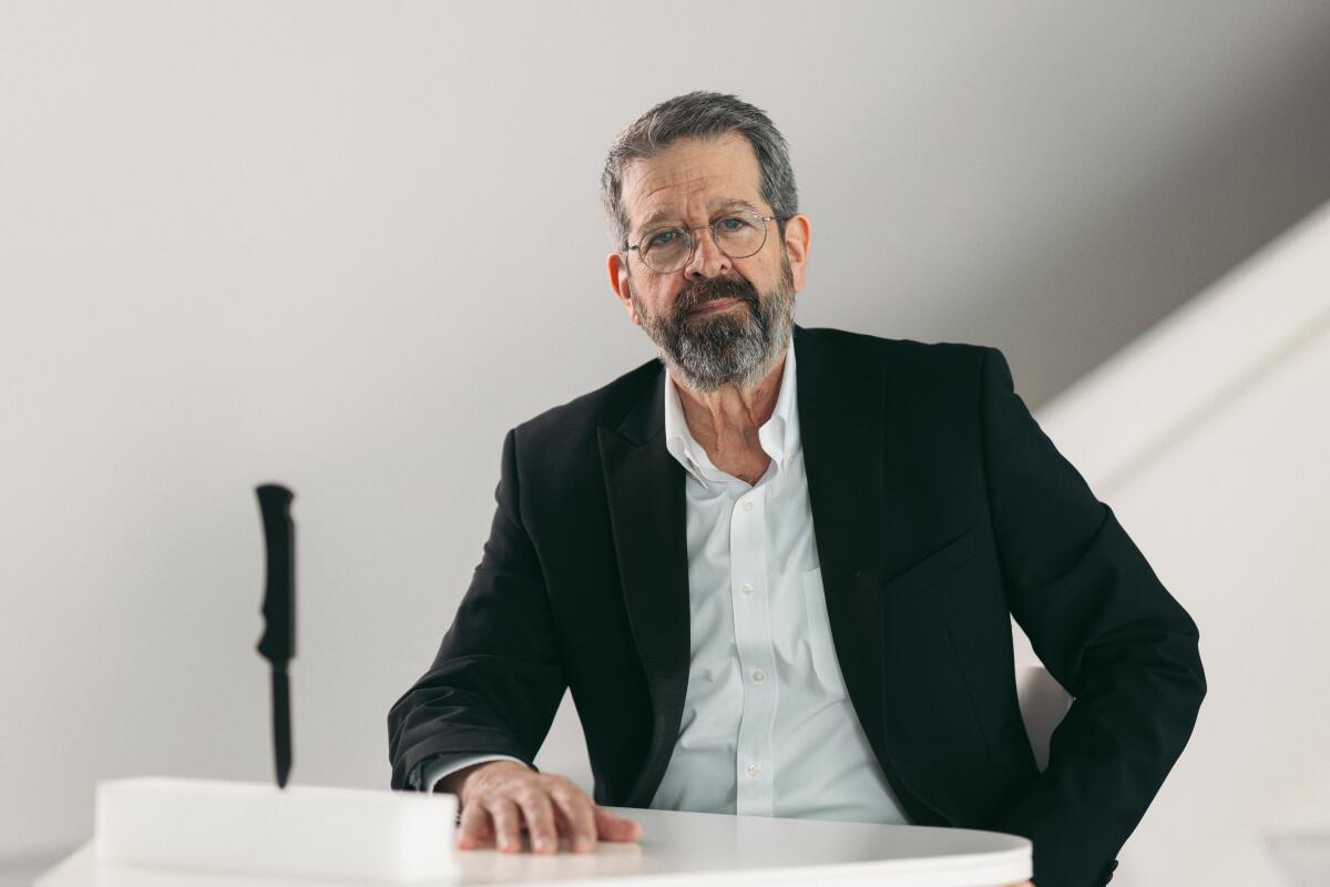 An actor poses with a knife embedded in a table next to him.