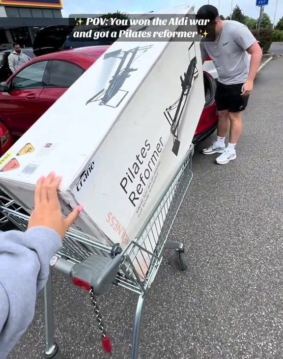 Person loading a Pilates reformer into a shopping cart outside an Aldi store.