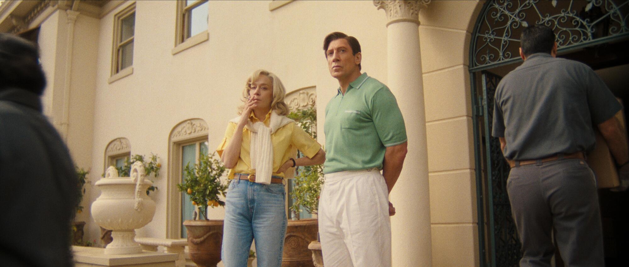 Two people stand outside a grand building as a moving man carries something past them