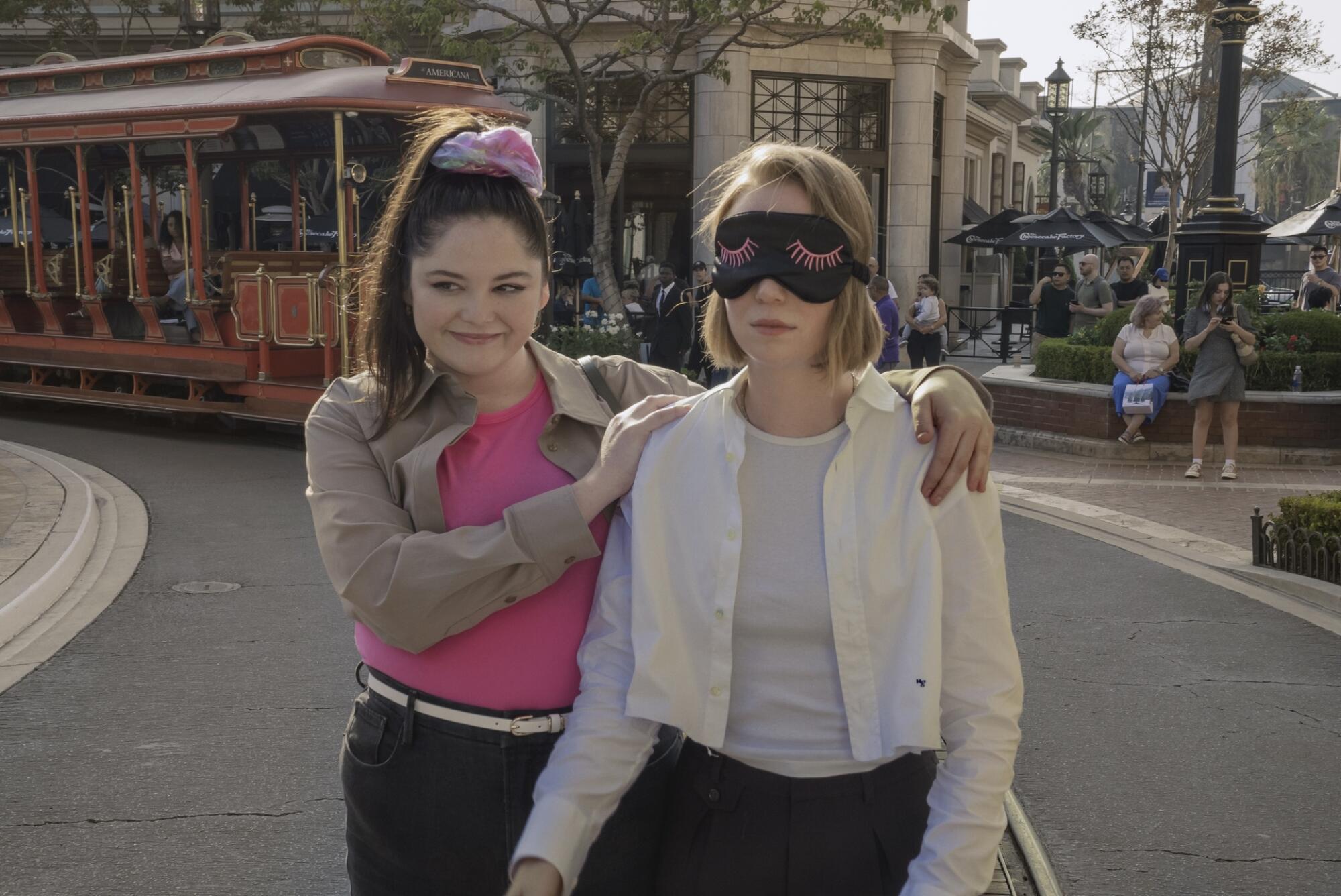 A woman walks a blindfolded woman down a paved road, a colorful trolley behind them
