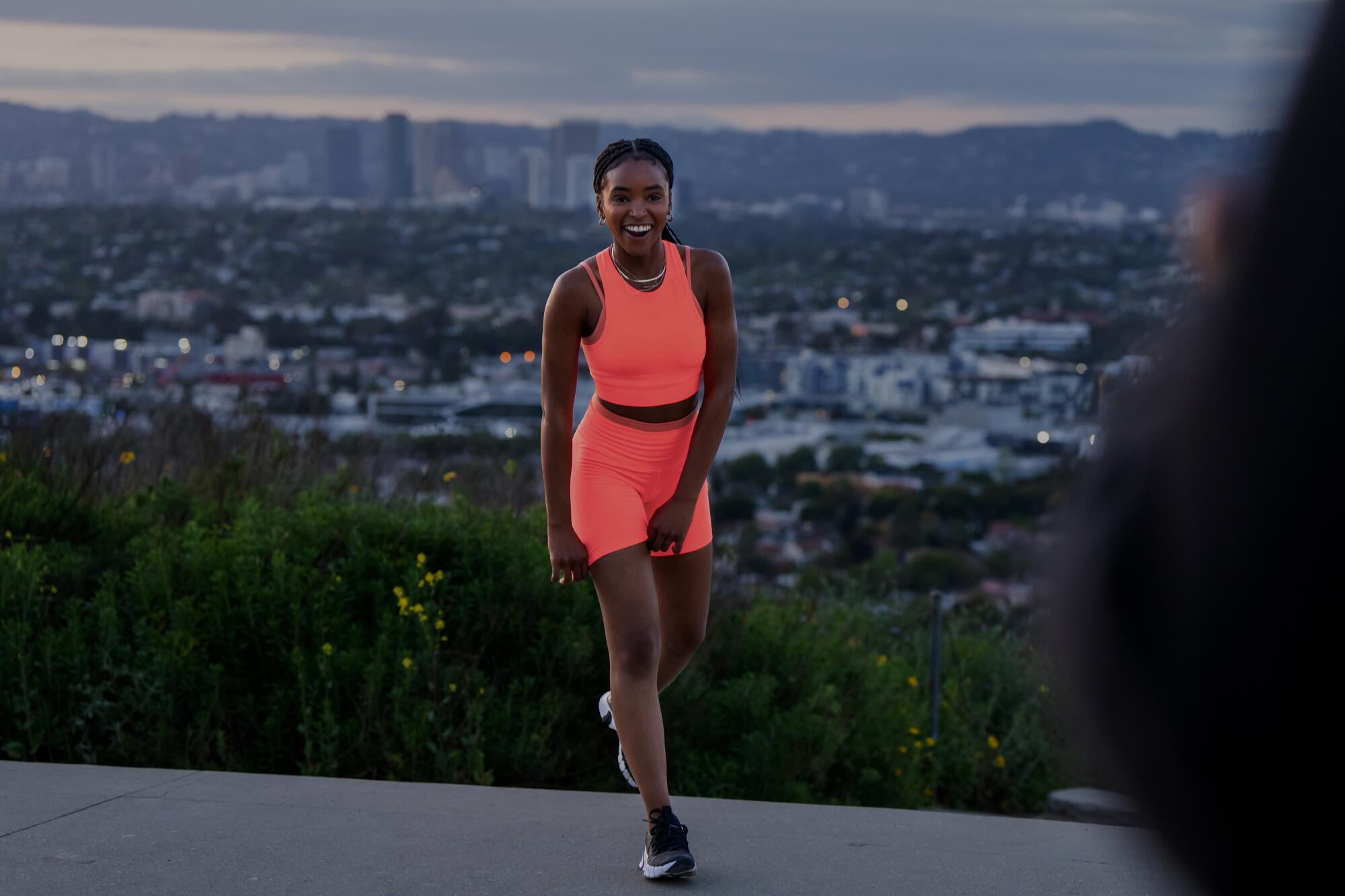 A woman in orange workout wear stands outside with a view of downtown Los Angeles behind her