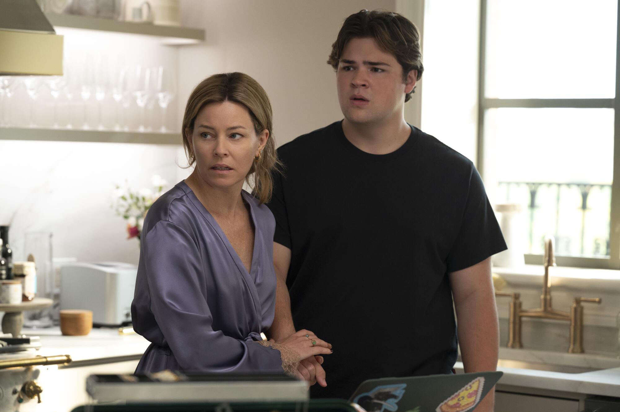 A woman in a purple silk robe holds the hand of a teenage boy in a black T-shirt. They are standing in a kitchen.