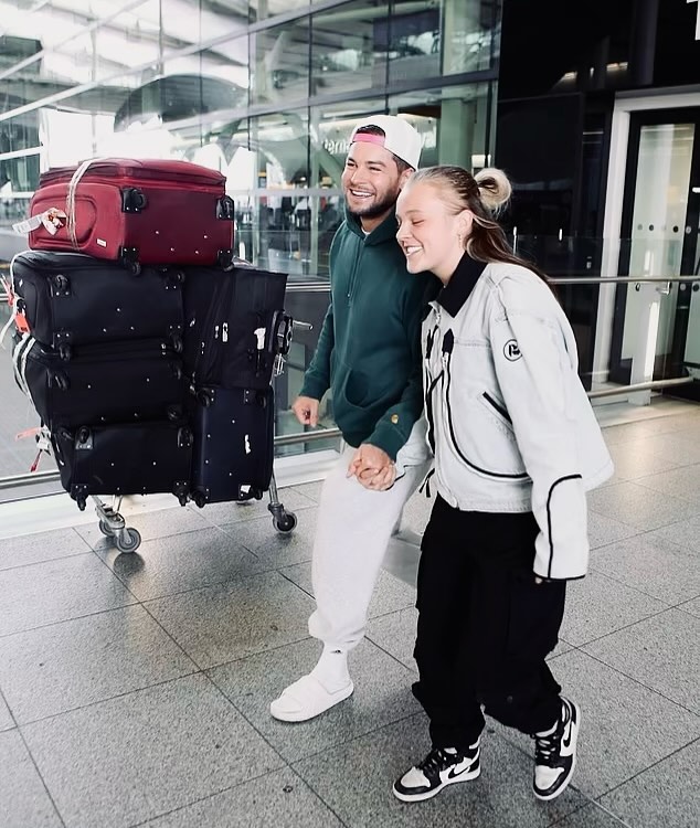 Jojo Siwa and Chris Hughes holding hands at an airport with luggage.