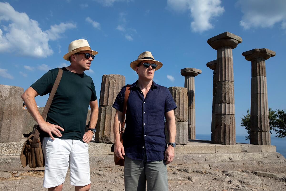 A man in white shorts, a greet T-shirt and hat stands next to a man in a blue shirt, gray pants and hat at a site in Greece.
