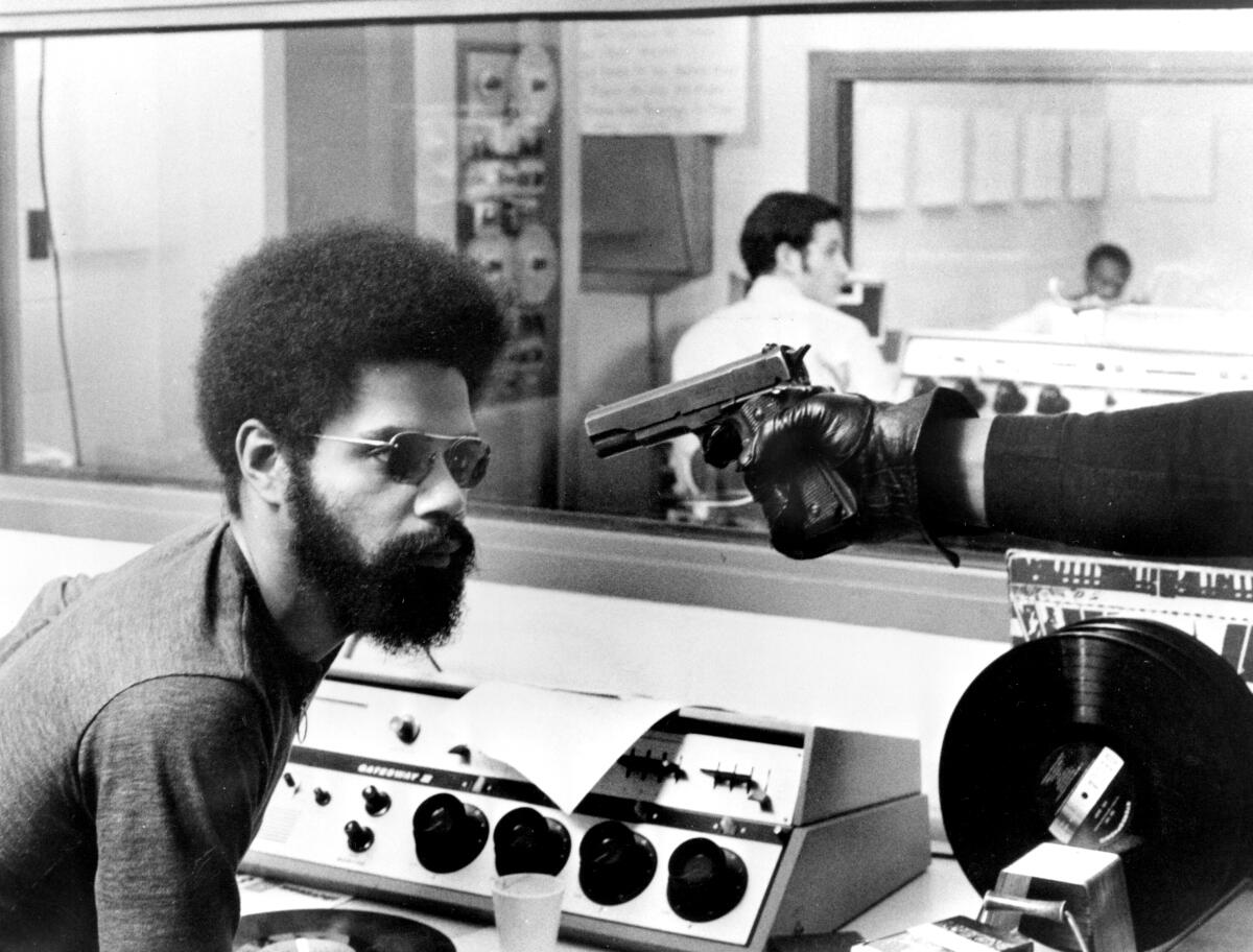 A man in an Afro sits by a recording console as a gun is pulled on him.