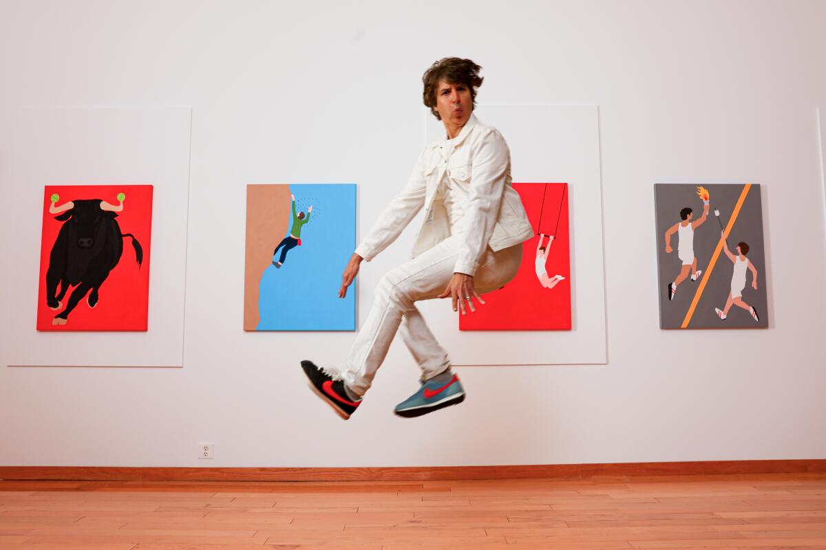 Demetri Martin, in a white suit, jumps in front of a white gallery wall with four colorful paintings.