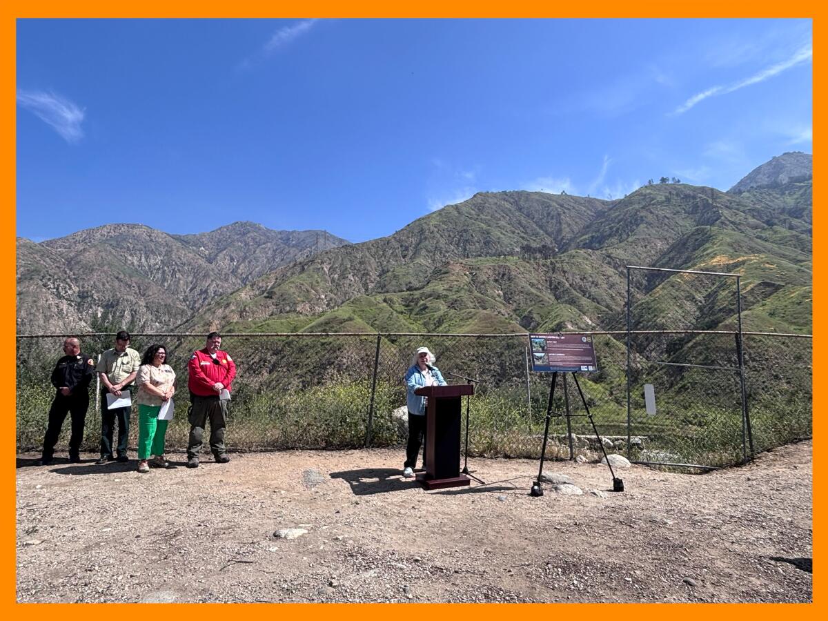 A person speaks during a news conference with other officials in the background near a fence.