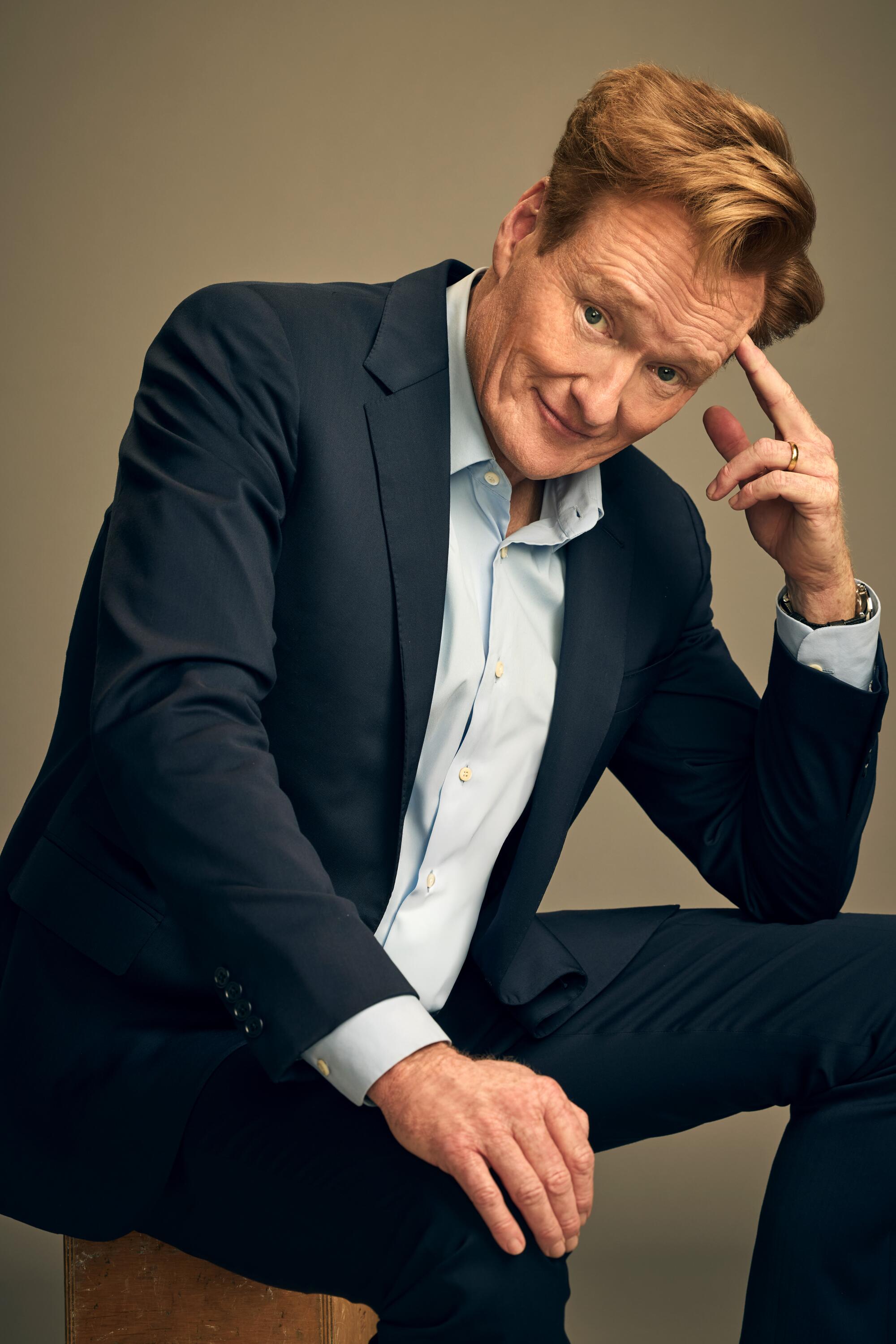 A man in a dark blue suit and light blue shirt sits on a box with one