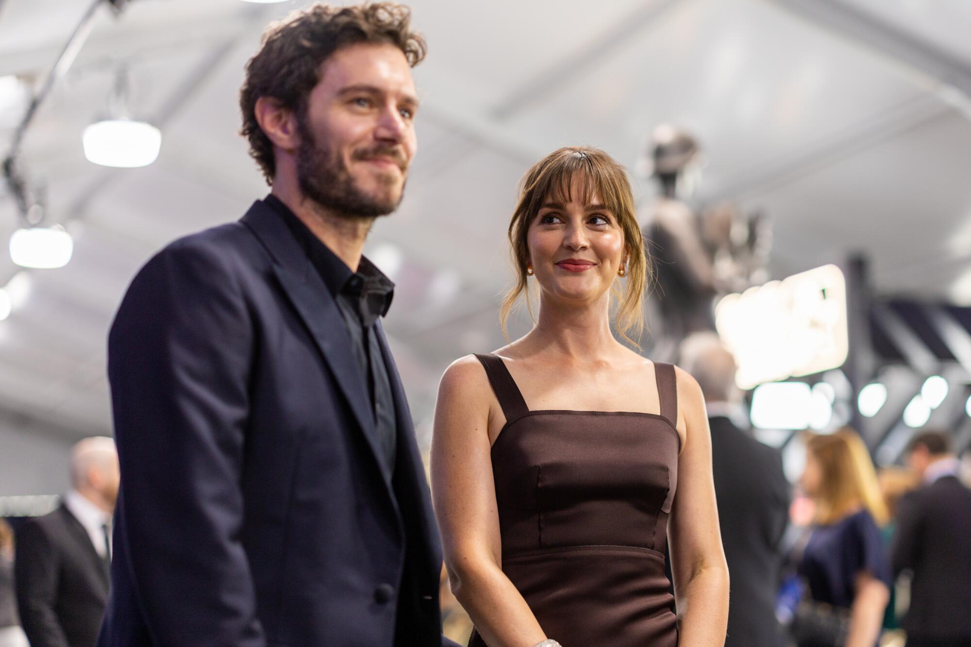 A bearded man in a suit smiles next to a smiling woman in a brown dress who's looking at him