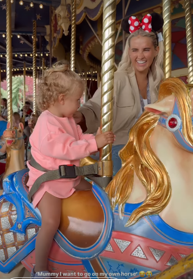 Toddler riding a carousel horse with her mother nearby.