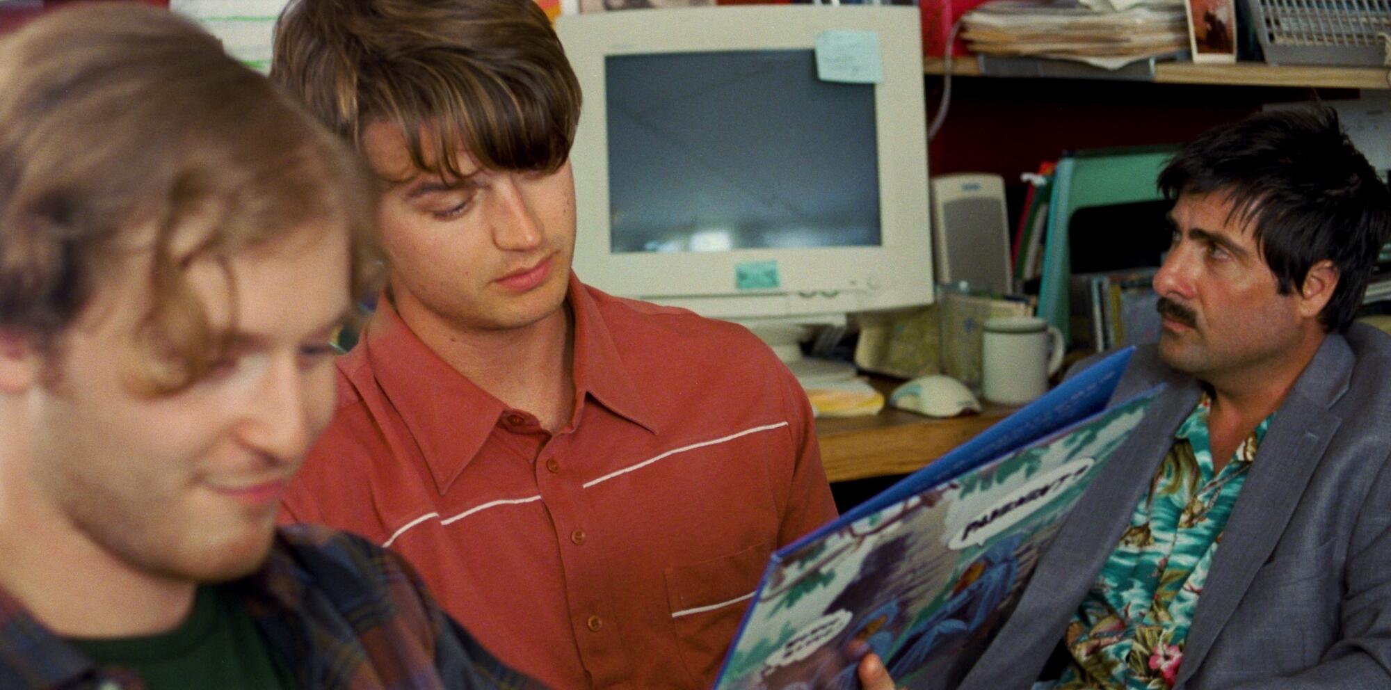 Band mates look at an album cover while a manager scowls.