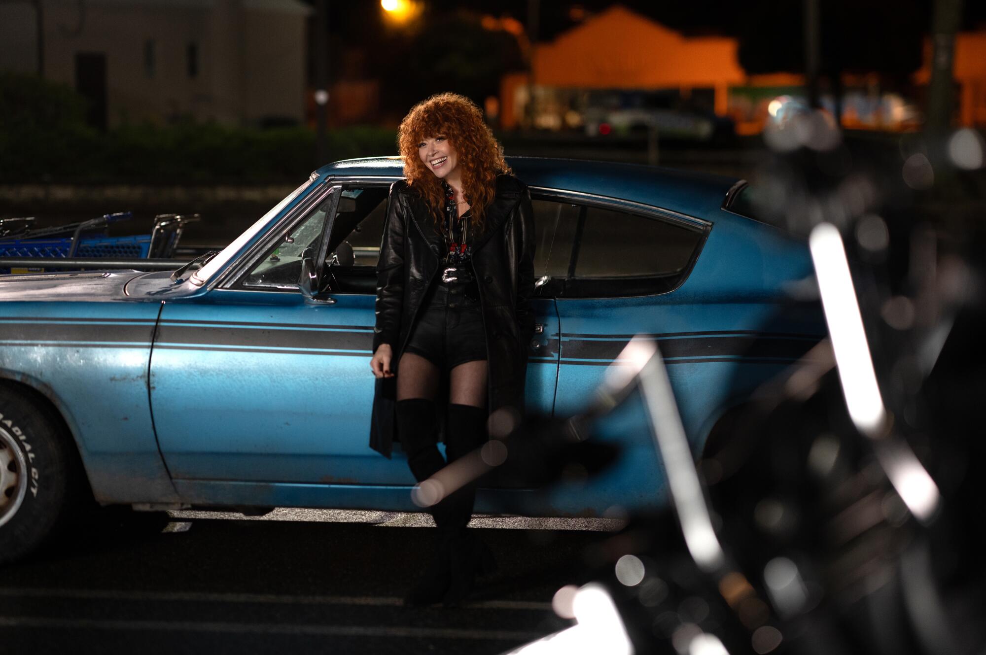 A woman in a black outfit leans against an older blue car with a black racing stripe.
