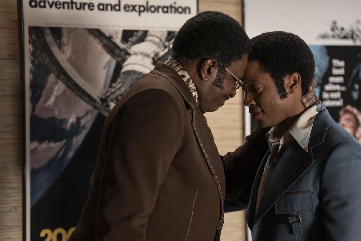 A man in a brown blazer leans his head onto a younger man in a blue leisure suit.