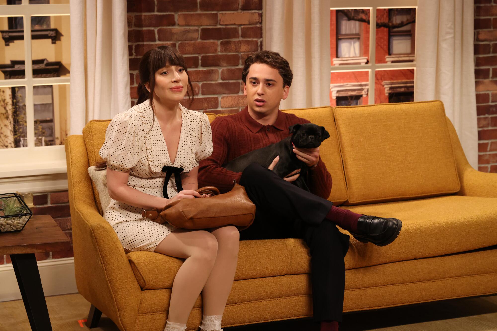 A woman in a white dress sits on a couch next to man in holding a small black dog.