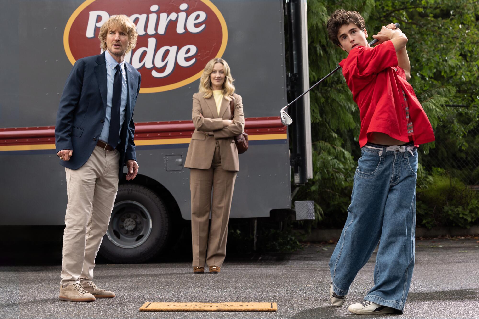 Two older adults stand near a box truck as a teenage boy in a red shirt and wide-leg jeans swings a golf club.