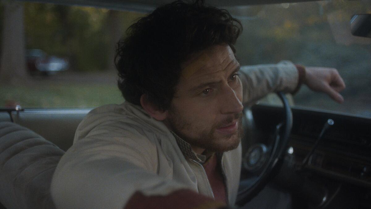 A man in a white jacket leans forward in a car nervously.