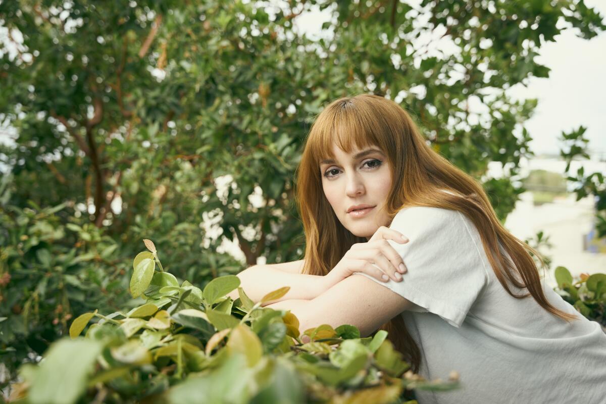 A woman with long red hair and bangs in a white T-shirt leans against a railing covered in green vines.